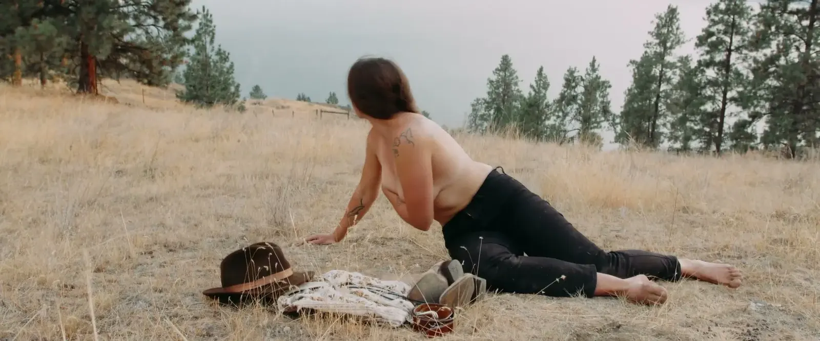 A woman lies on dry grass beside a hat and folded shirt.