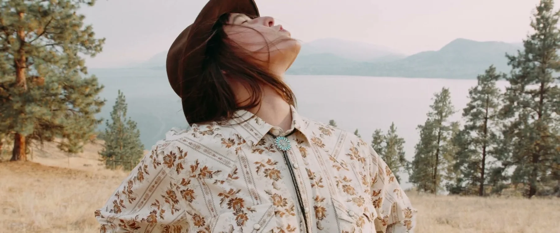 A woman in a wide-brimmed hat looks upward above a lake.
