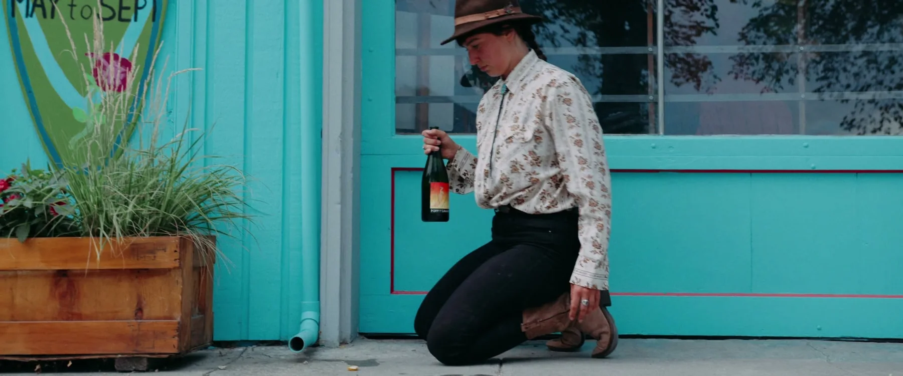 A woman kneels with a bottle outside a turquoise building.