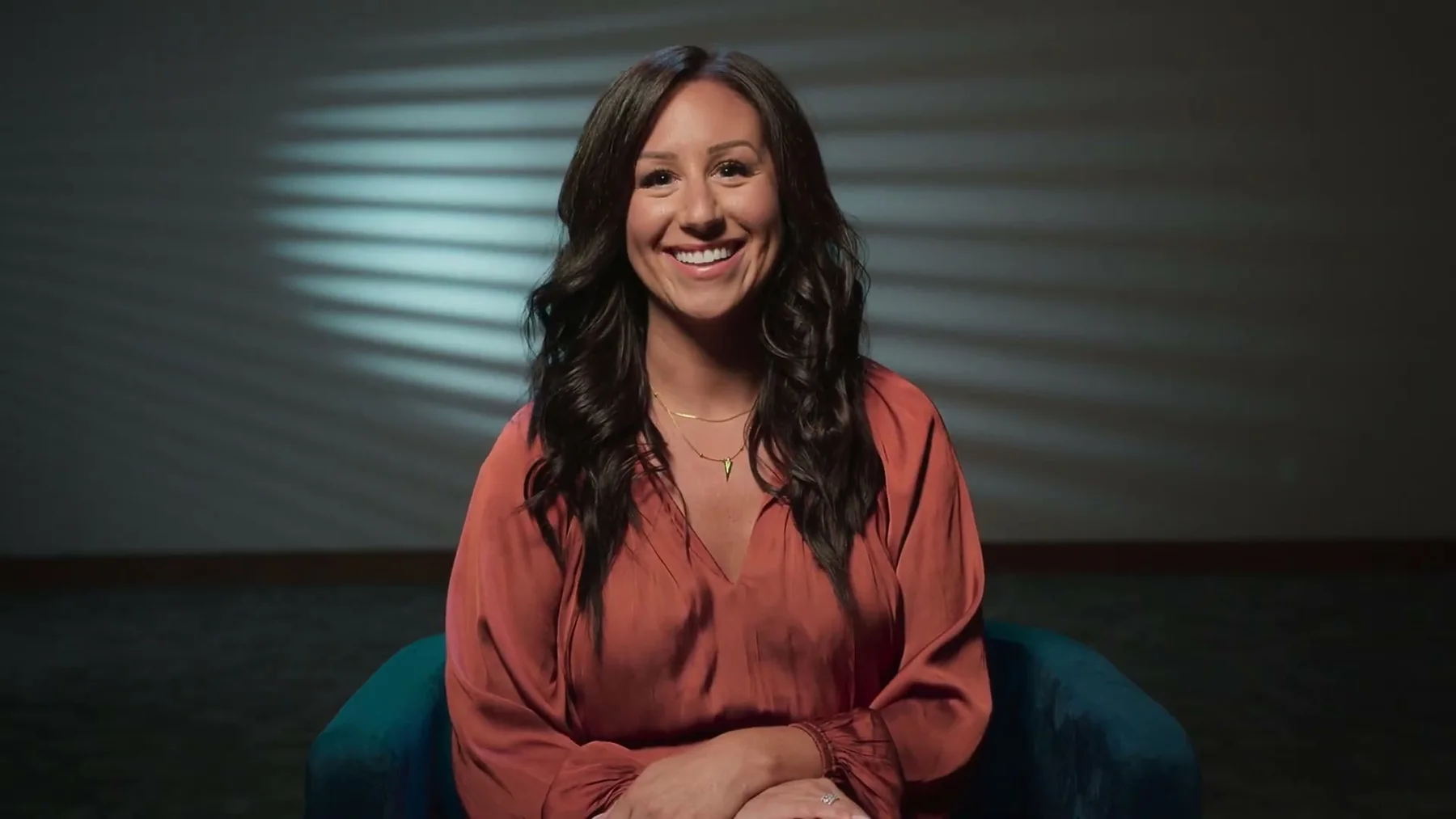 A woman sits for an interview against a dark studio background.