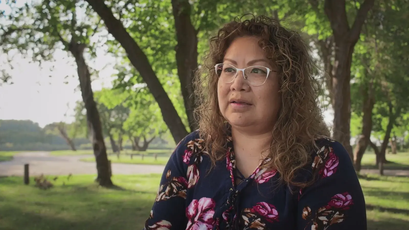A woman sits for an interview beneath trees in a park.