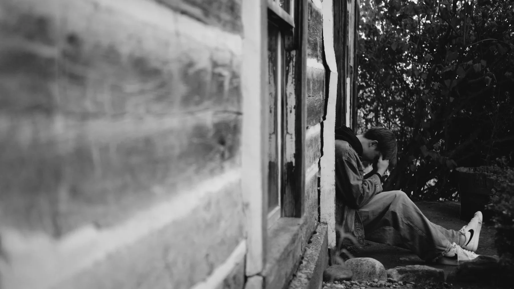 A man sits in the doorway of a weathered vehicle in black and white.
