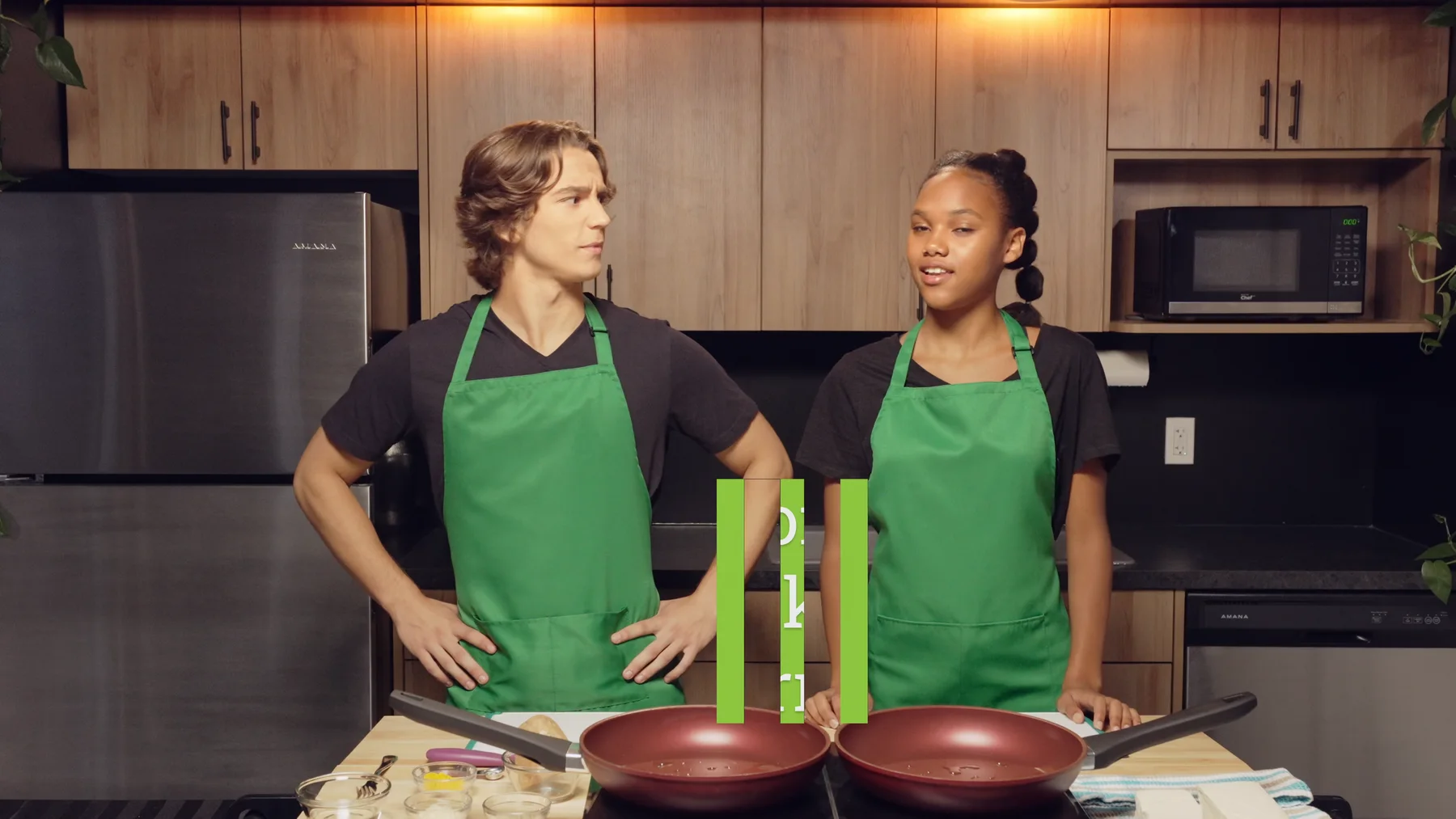 Two EcoCooks hosts in green aprons standing at a kitchen counter during a kids cooking-show video.