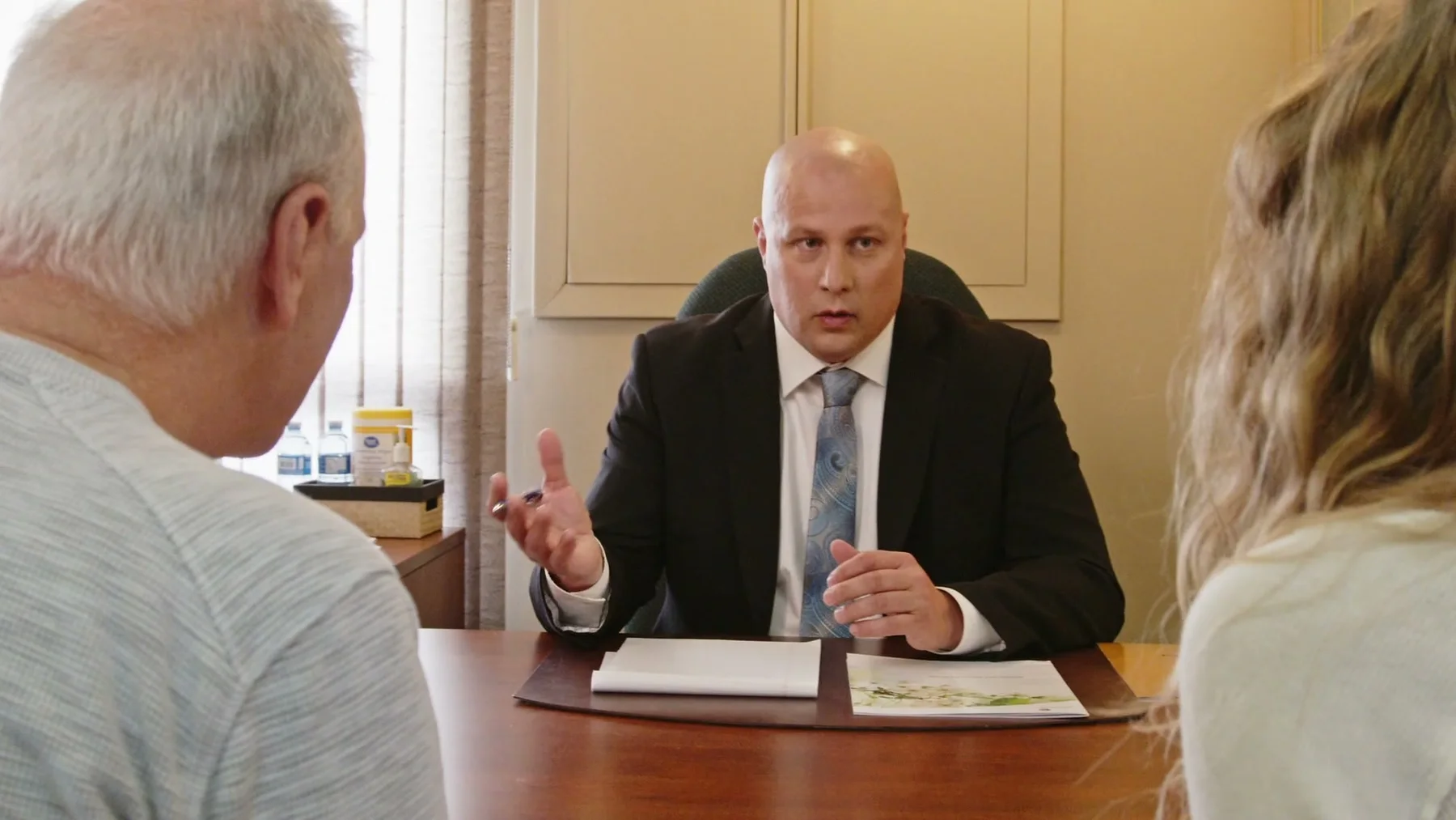 A man in a suit speaks with two people across an office desk.