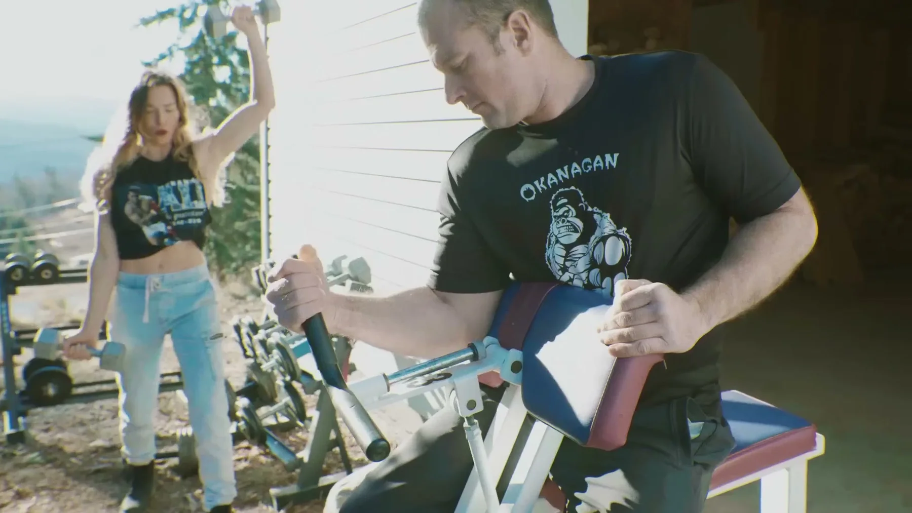 A man grips an arm-wrestling trainer while a woman lifts dumbbells outside.