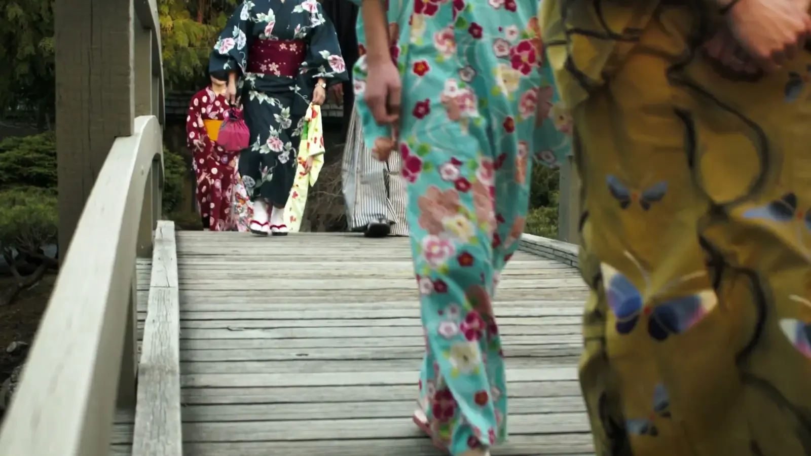 Women in colourful kimono walk across a wooden garden bridge.