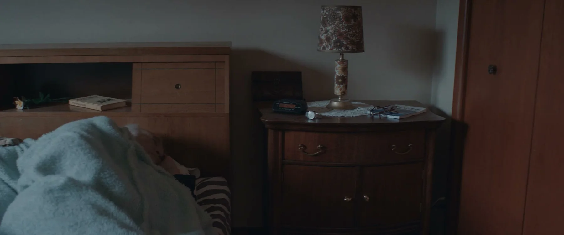 A dim bedroom with a bed, wooden dresser and bedside lamp.