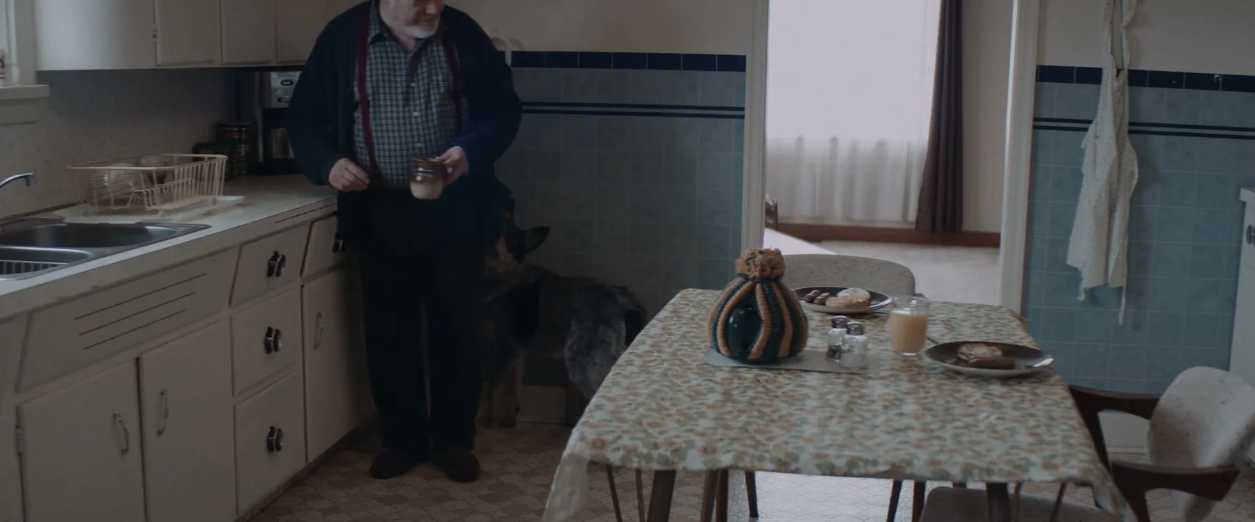 An older woman stands beside a kitchen table set with food.