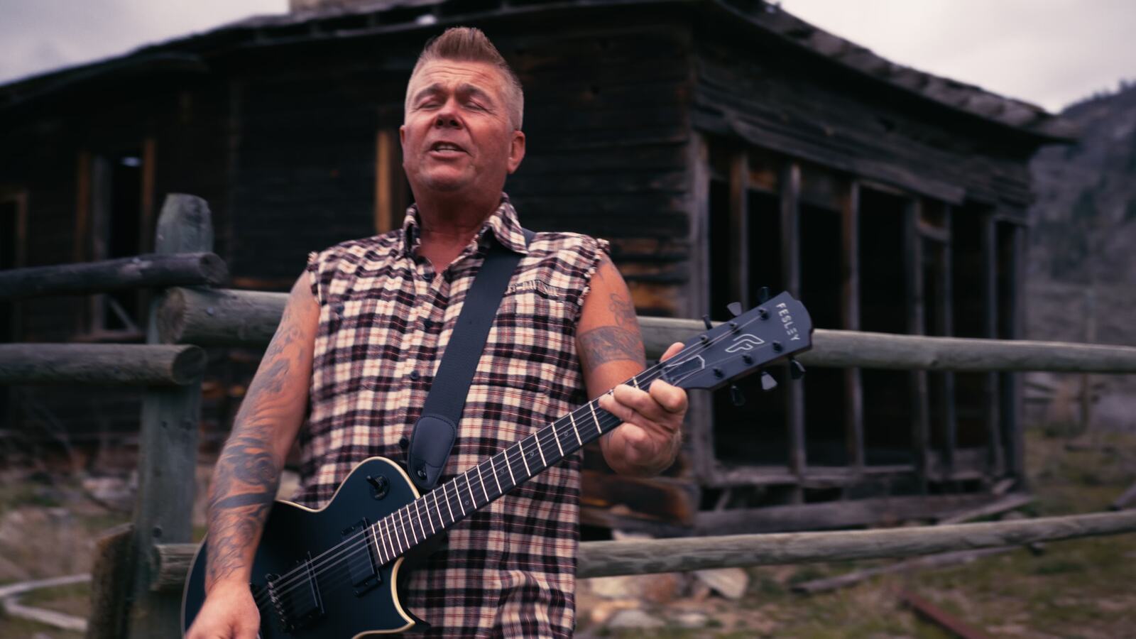 Trevor Ingram performs with a guitar in front of a weathered wooden building.