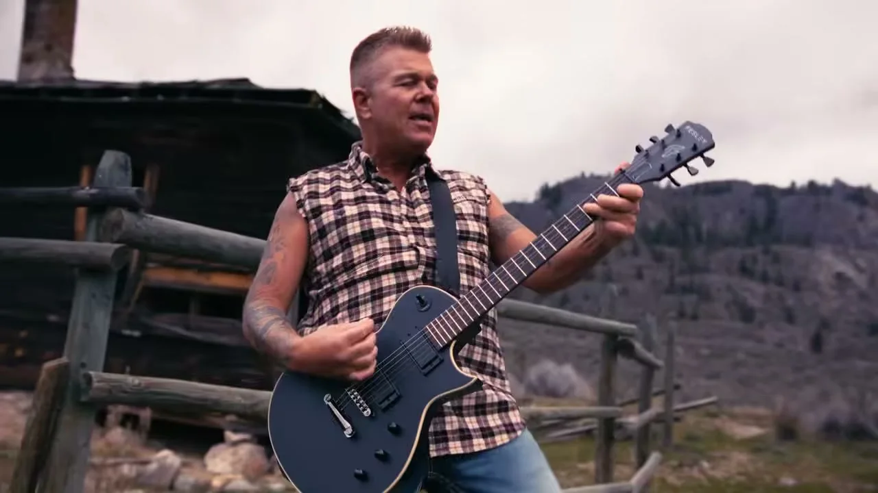 Trevor Ingram plays an electric guitar beside a wooden cabin.