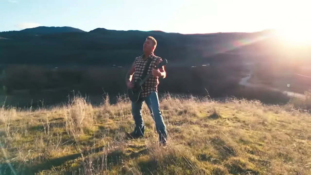 Trevor Ingram performs with a guitar on a sunlit hillside.