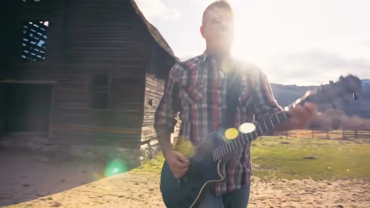 Trevor Ingram plays guitar beside a weathered wooden building.