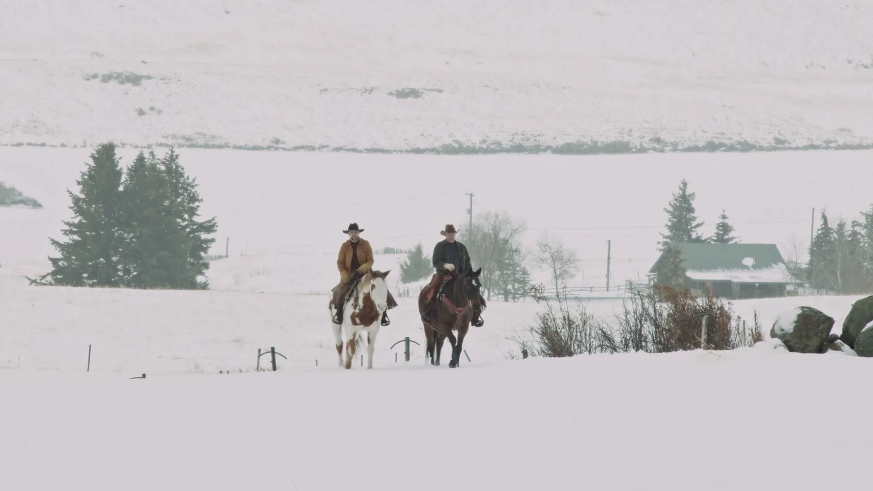 Two riders cross a snowy ranch field on horseback.
