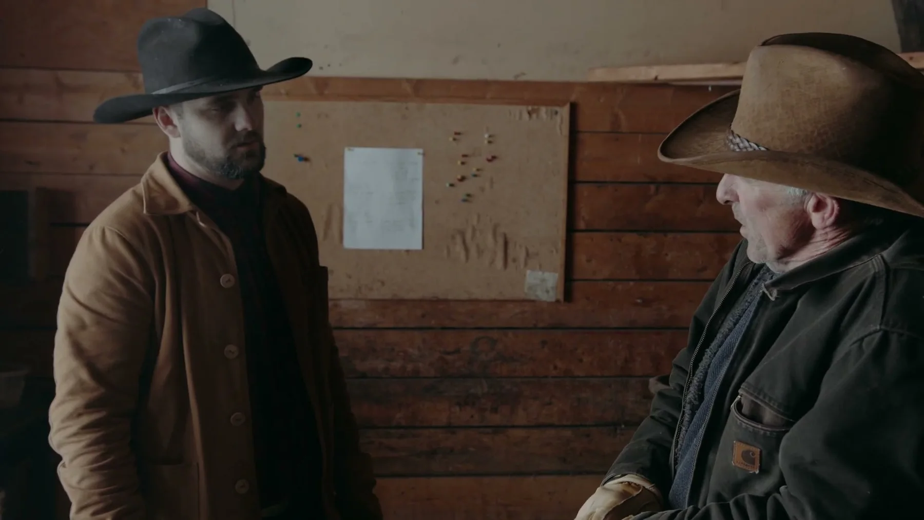 Two men in cowboy hats face one another inside a wooden room.