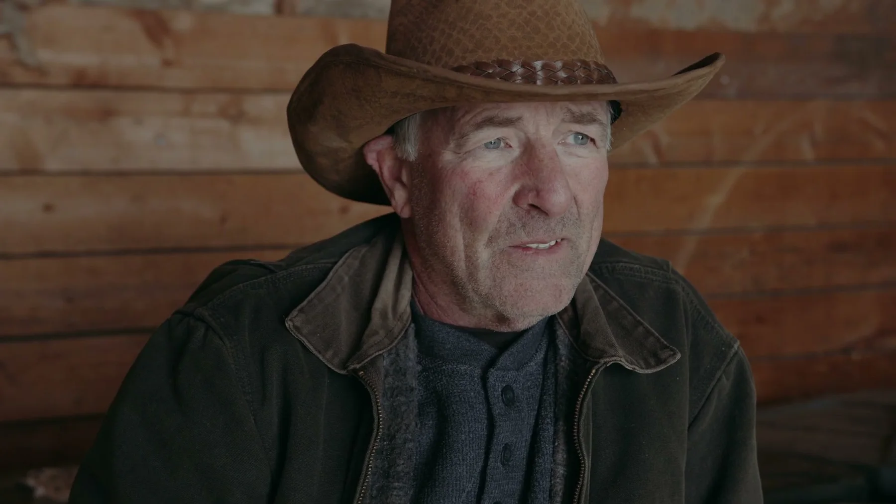 An older man in a brown cowboy hat stands against a wooden wall.