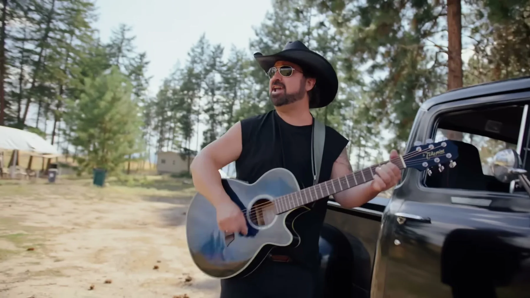 Kris Anders plays guitar beside a black pickup truck in a forest clearing.