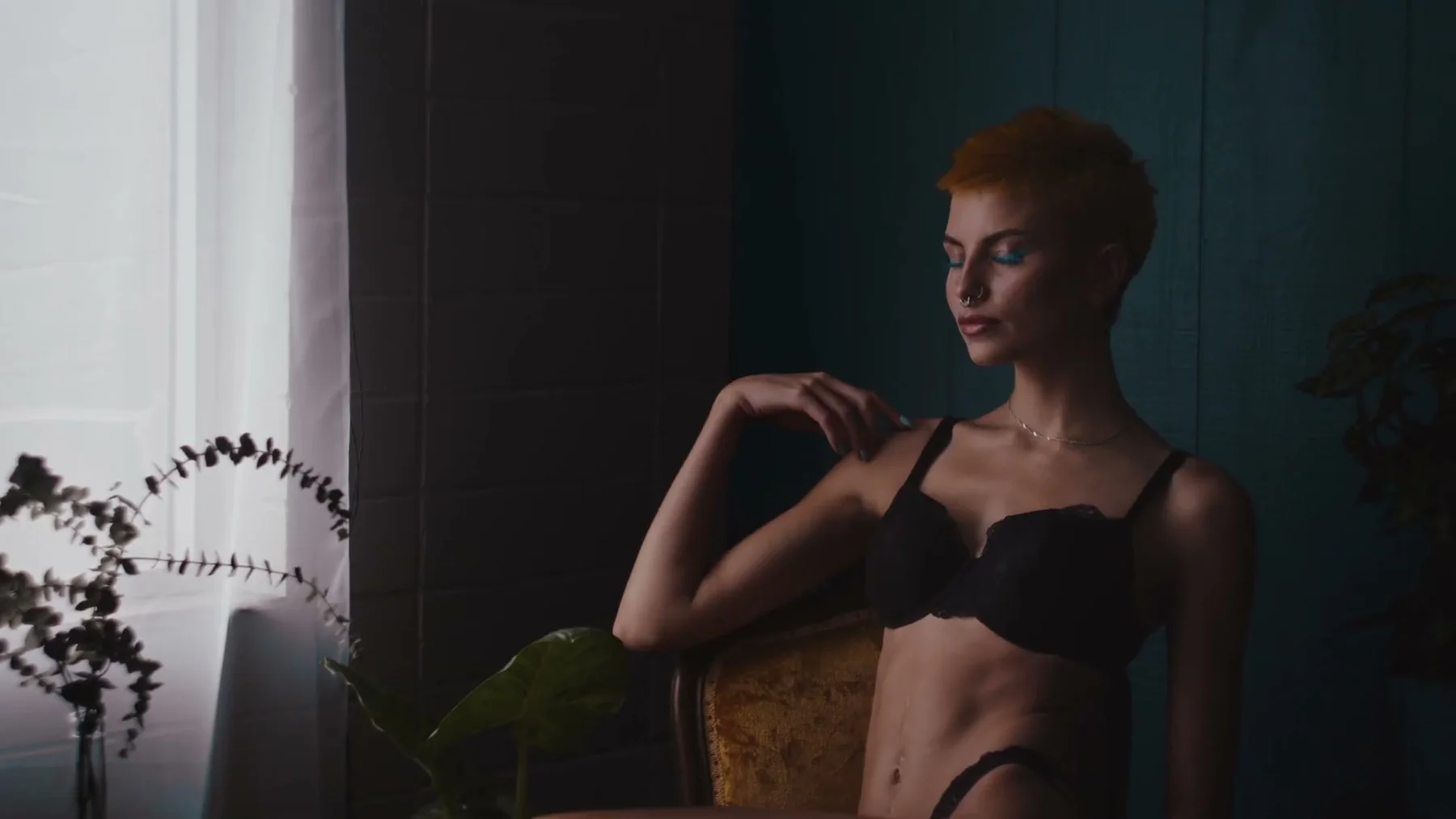 A model stands beside a window in black lingerie against a teal wall.