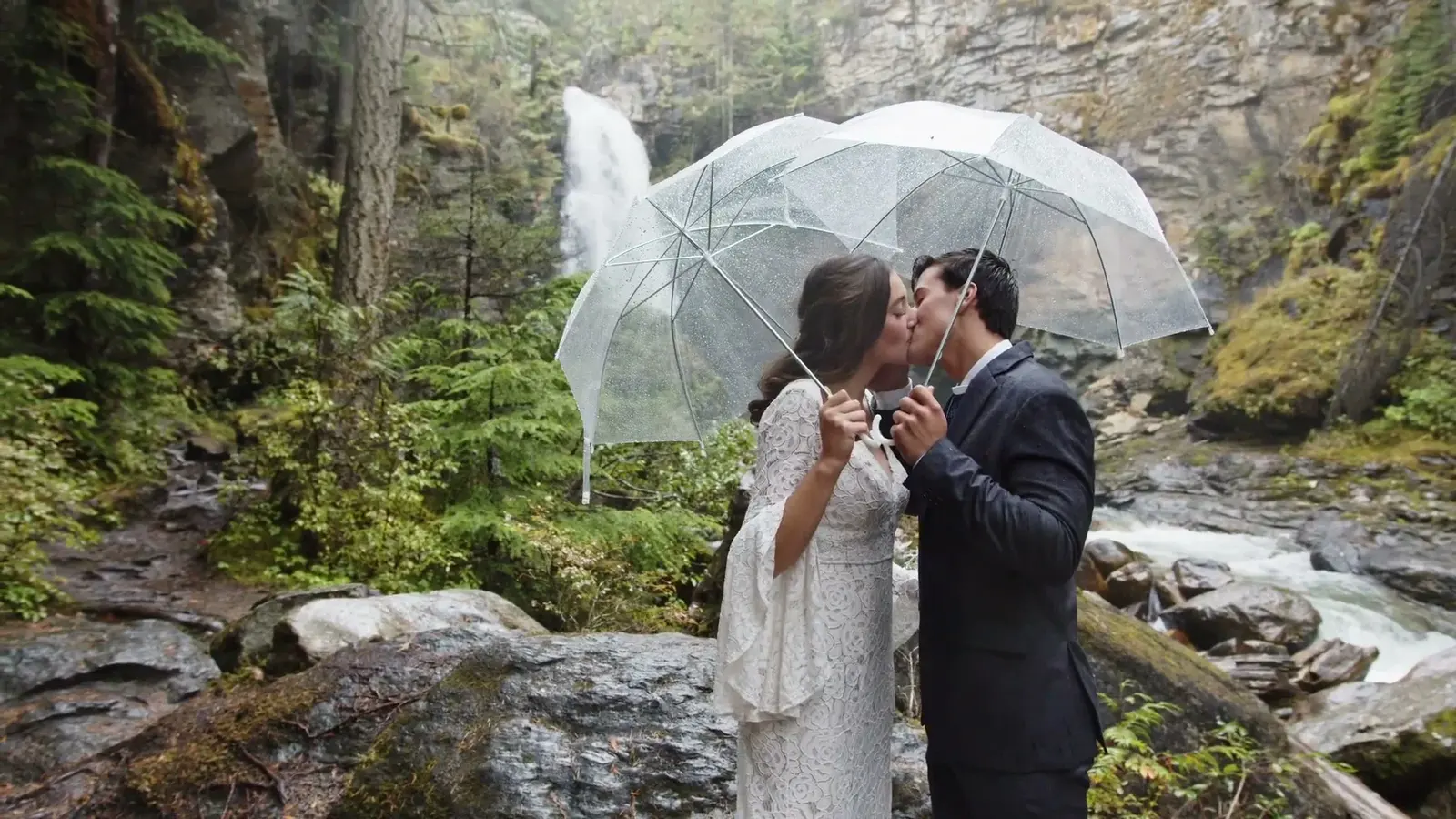 A couple stands beneath clear umbrellas beside a waterfall.