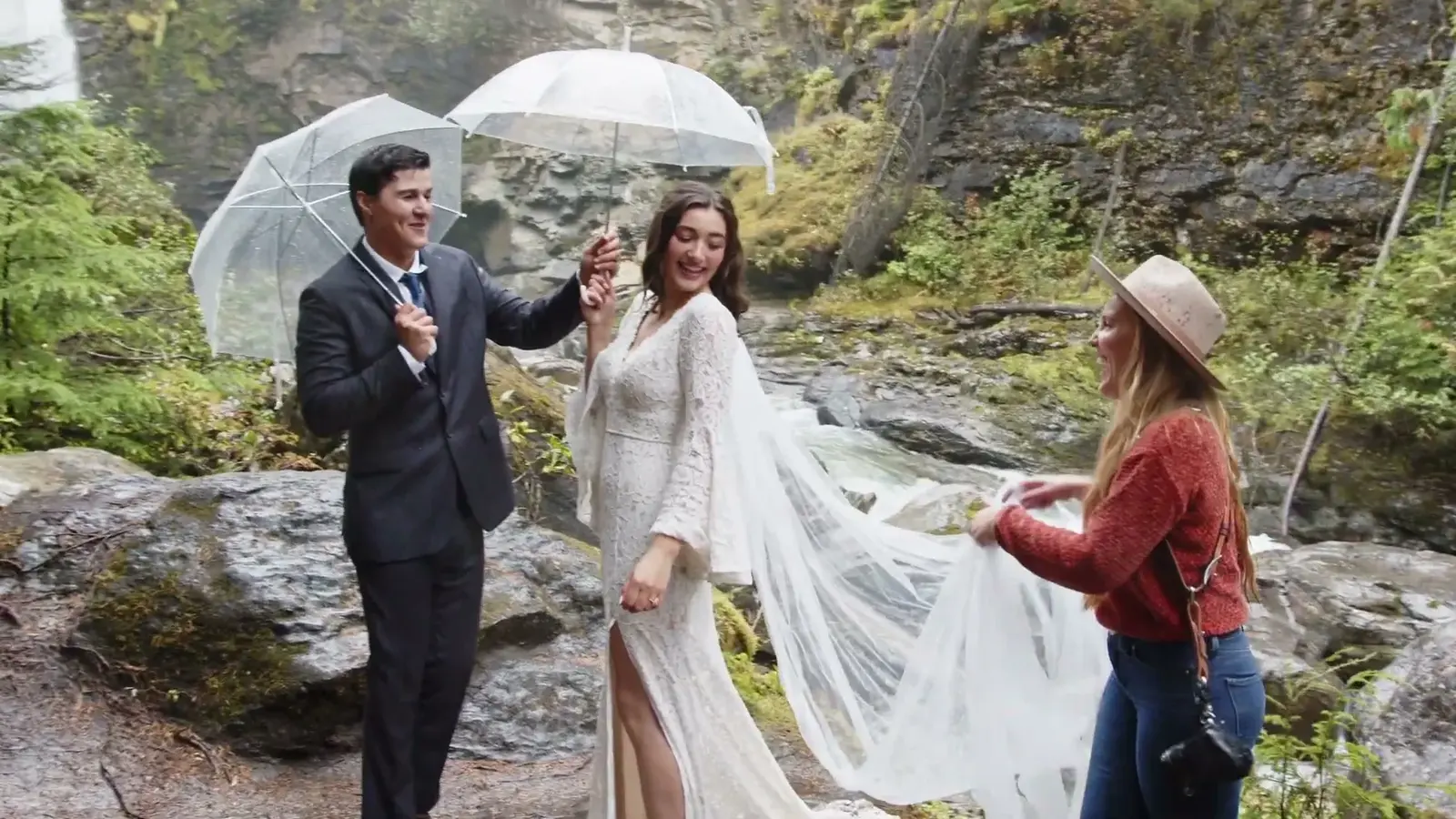 A photographer adjusts a bride's veil beside a waterfall.