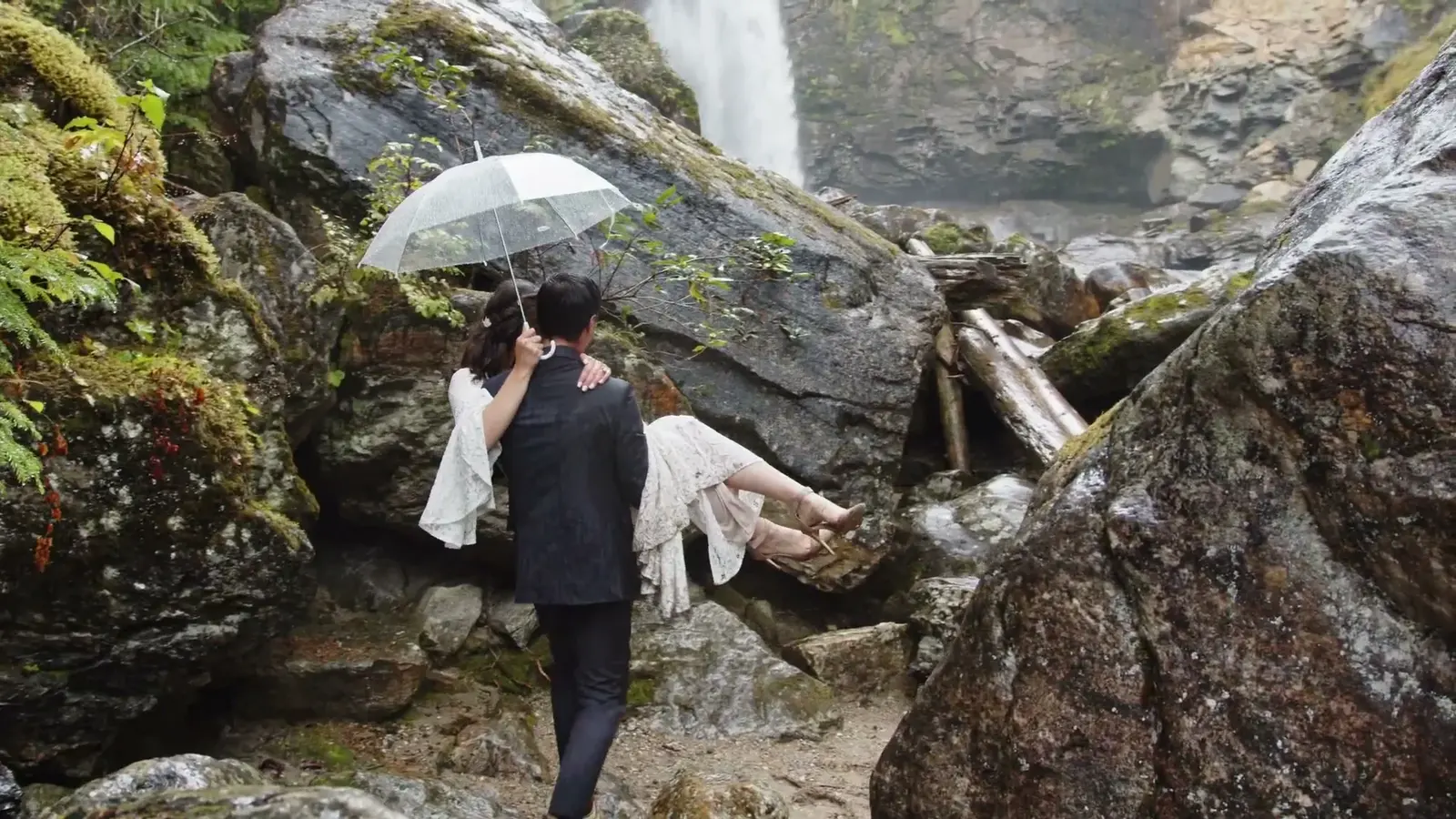 A couple walks across rocks beneath an umbrella toward a waterfall.