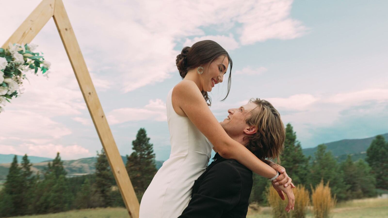 A bride laughs as her partner lifts her beneath a wooden wedding arch.
