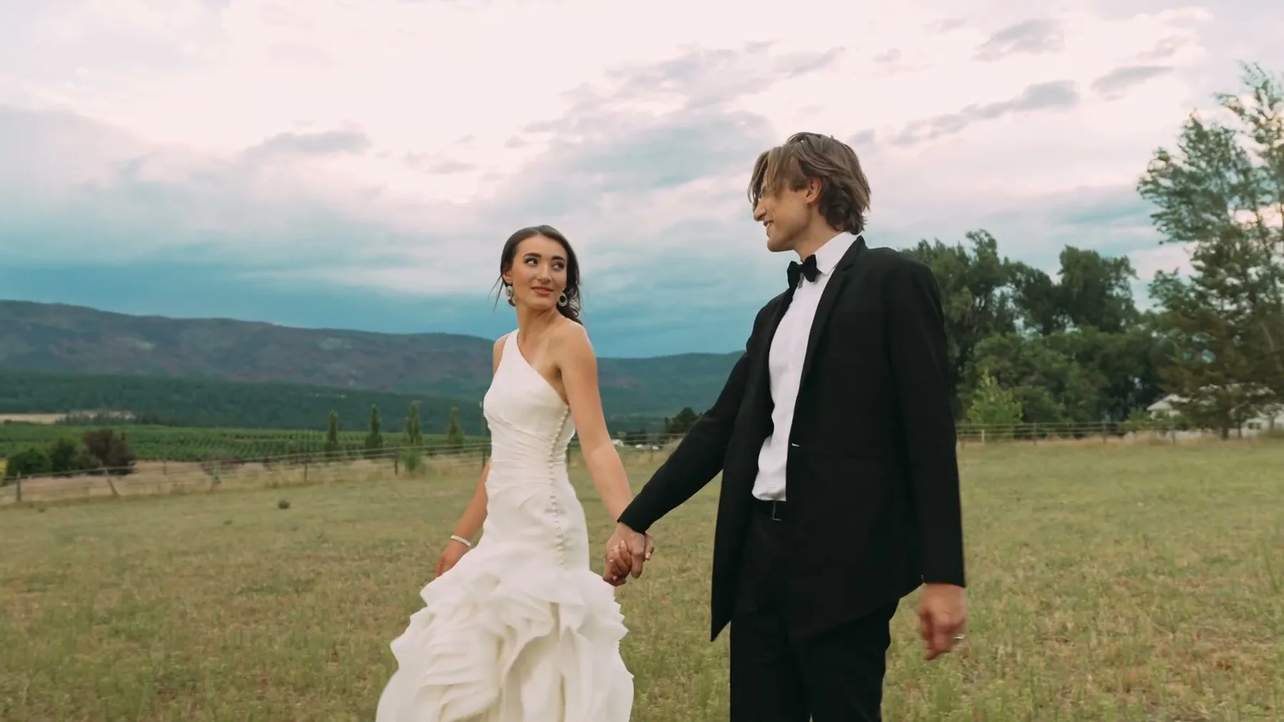 A bride and groom walk hand in hand through a ranch field.