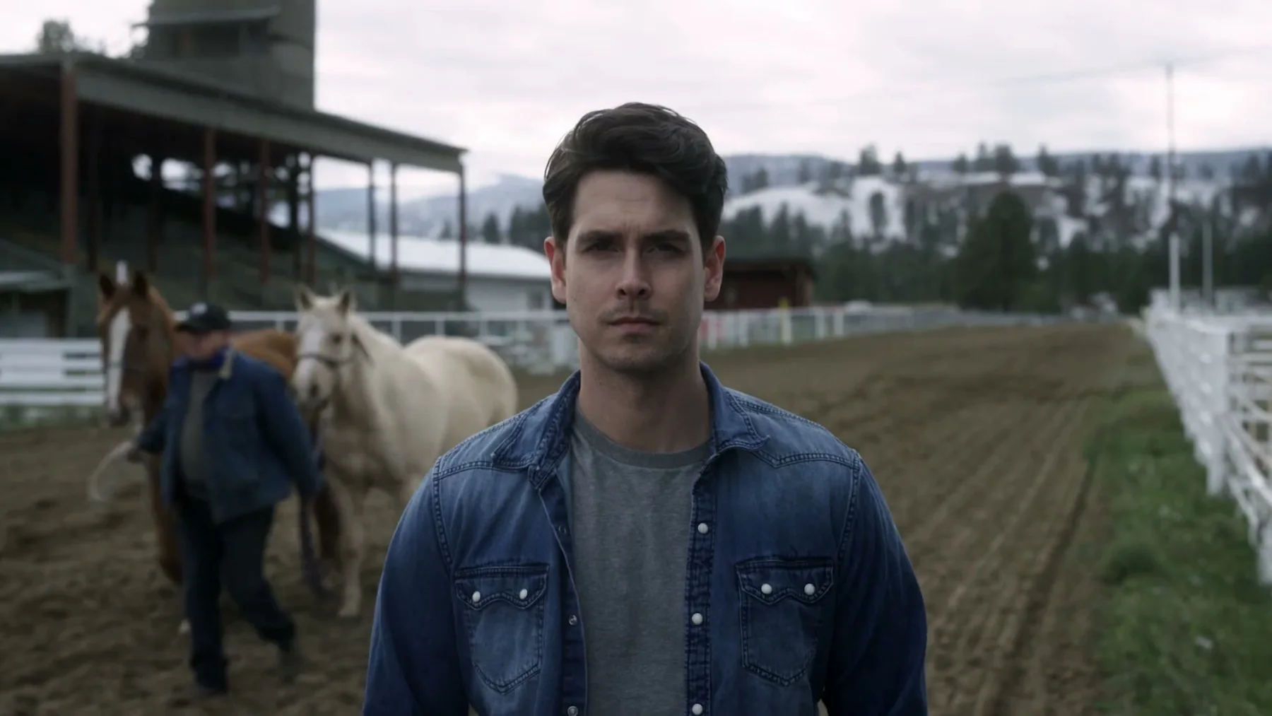 A man stands beside two horses in a fenced riding area.