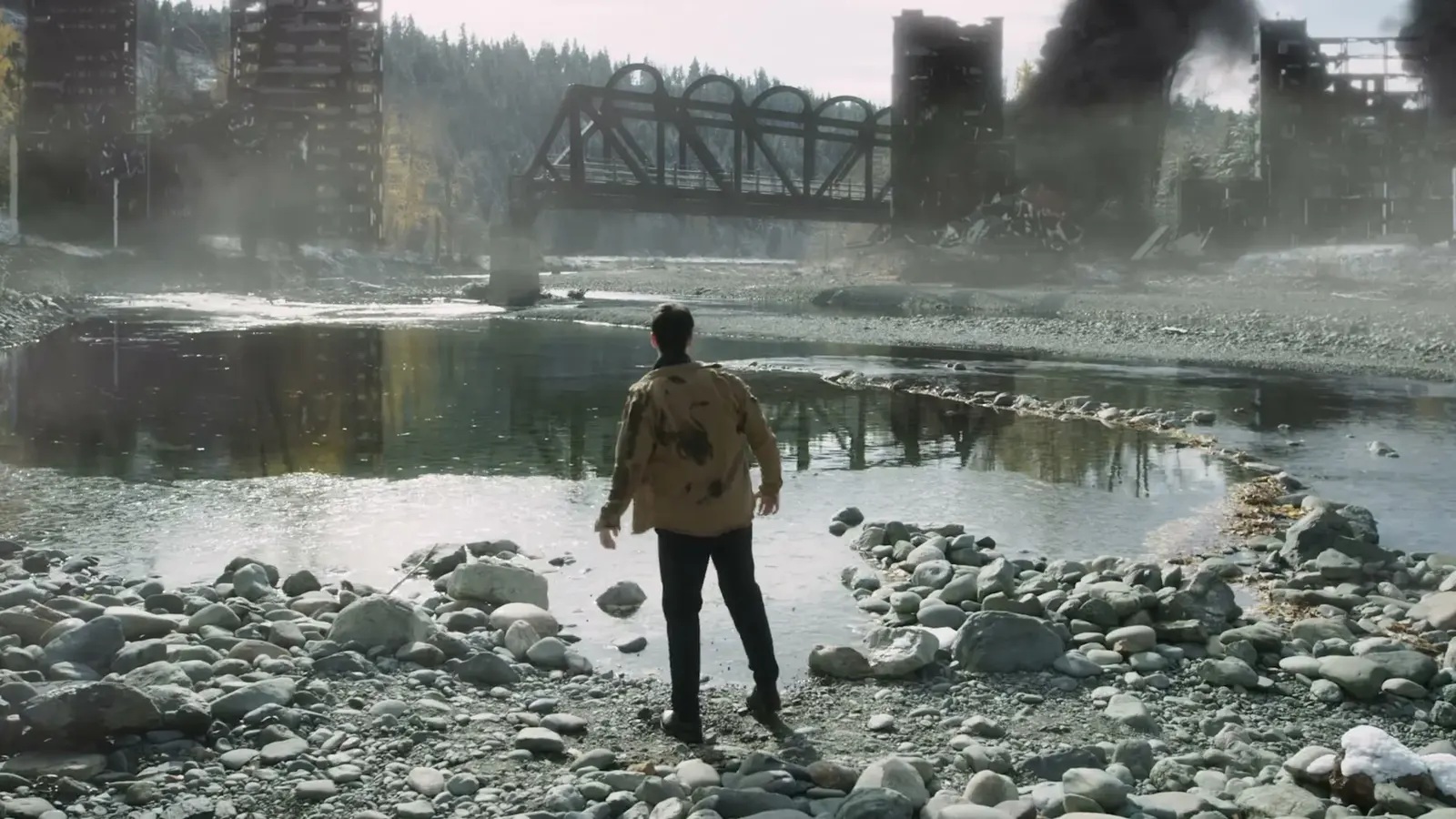 A man stands on a rocky riverbank near a metal bridge.